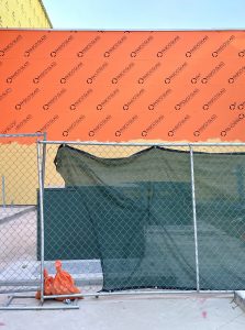 Construction site with an orange wall in the distance, a fence covered partially in black cloth in the foreground, and metal boxes in between.