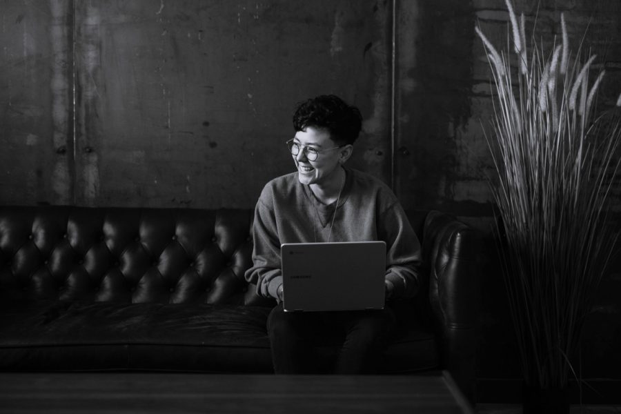 A black and white photo where a successful writer types at a computer next to a tall plant while sitting on a couch.