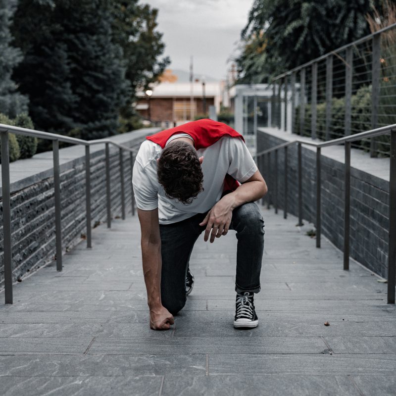 Man wearing a red cape over his clothes crouches with his fist punching the ground