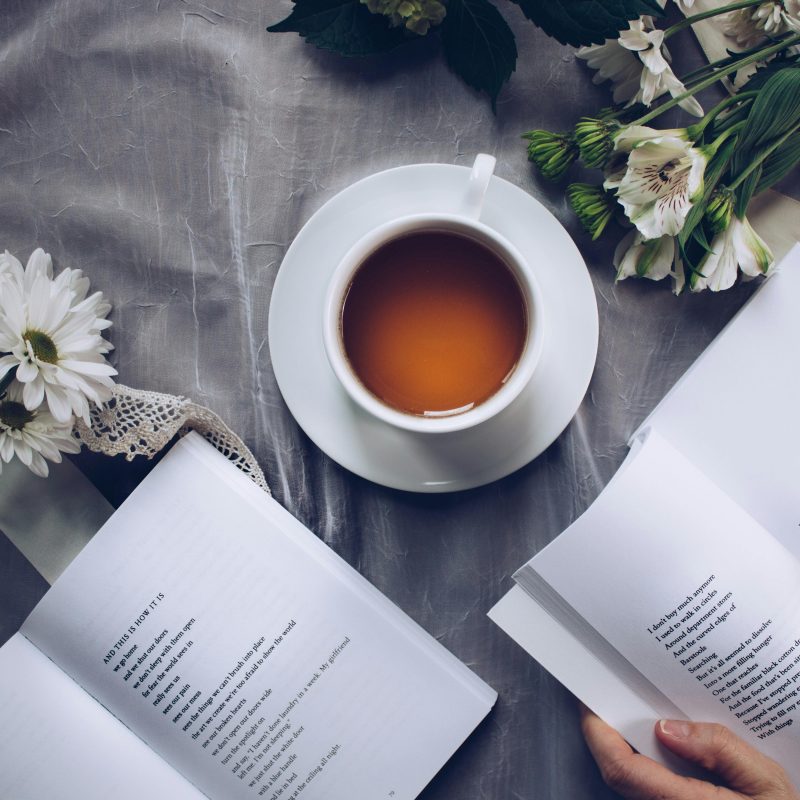 An above view of a table, on which sits a mug of tea, some flowers, and two books, one of which is being opened by a pair of hands.