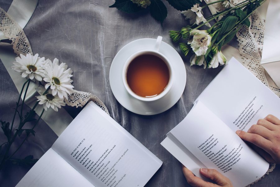 An above view of a table, on which sits a mug of tea, some flowers, and two books, one of which is being opened by a pair of hands.