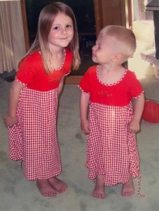 Two young children, a brother and sister, in matching red dresses. The little boy is smiling at his sister, and the little girl is smiling at the camera.